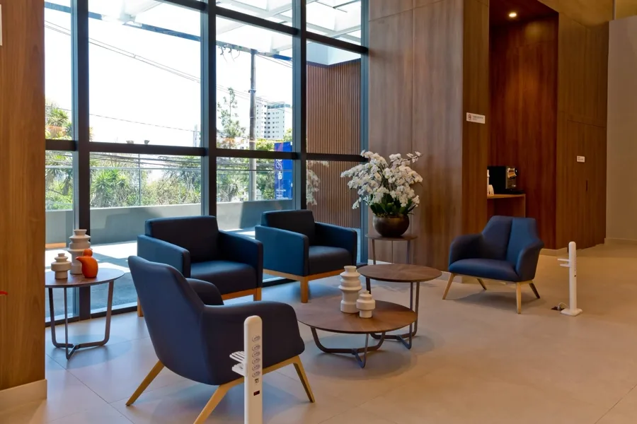 Lobby do hospital com poltronas azuis, pé-direito alto e fachada envidraçada