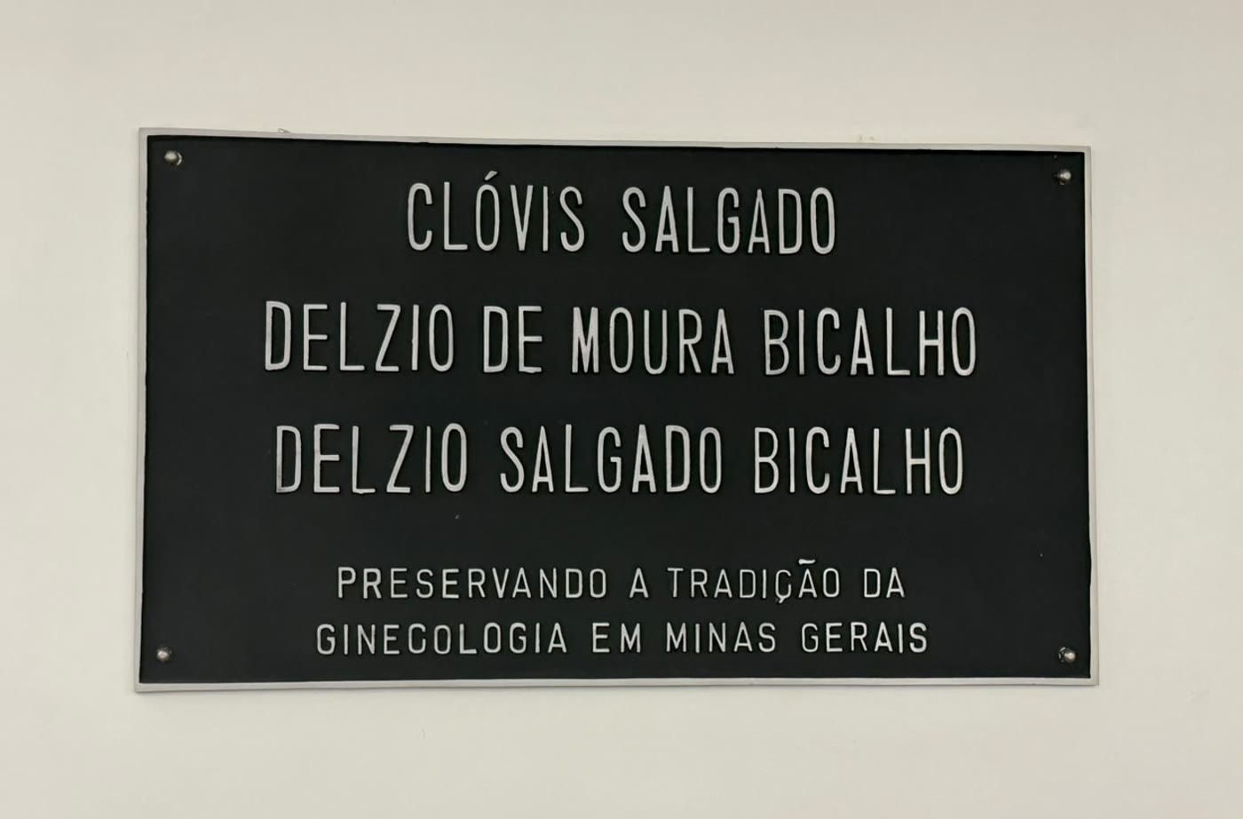 Placa na parede do consultório com os nomes de Clóvis Salgado, Delzio de Moura Bicalho e Delzio Salgado Bicalho e a inscrição "preservando a tradição da ginecologia em Minas Gerais"