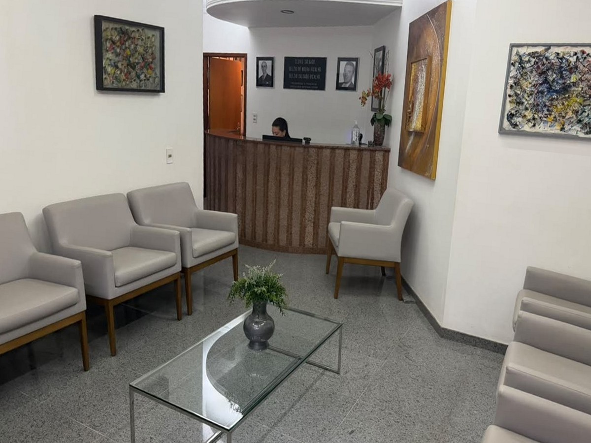 Sala de espera da clínica com poltronas claras, mesa de centro de vidro e o balcão de atendimento ao fundo